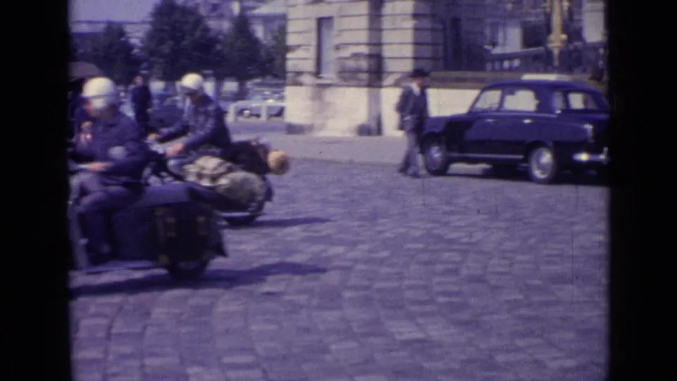 A scooter with two riders and luggage passes a dark compact car and pedestrians beside formal stone buildings in 1962 Versailles.