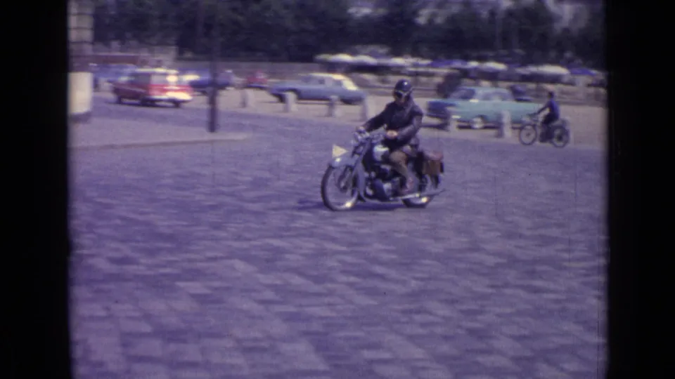 A lone motorcyclist crosses an open cobblestone area in Versailles, with mid-century cars and distant cyclists behind him.