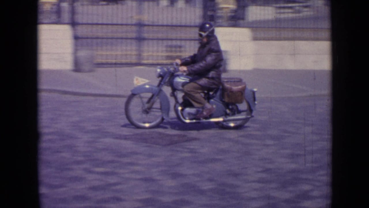 Helmeted rider on a motorcycle passing in front of a stone facade and fence.