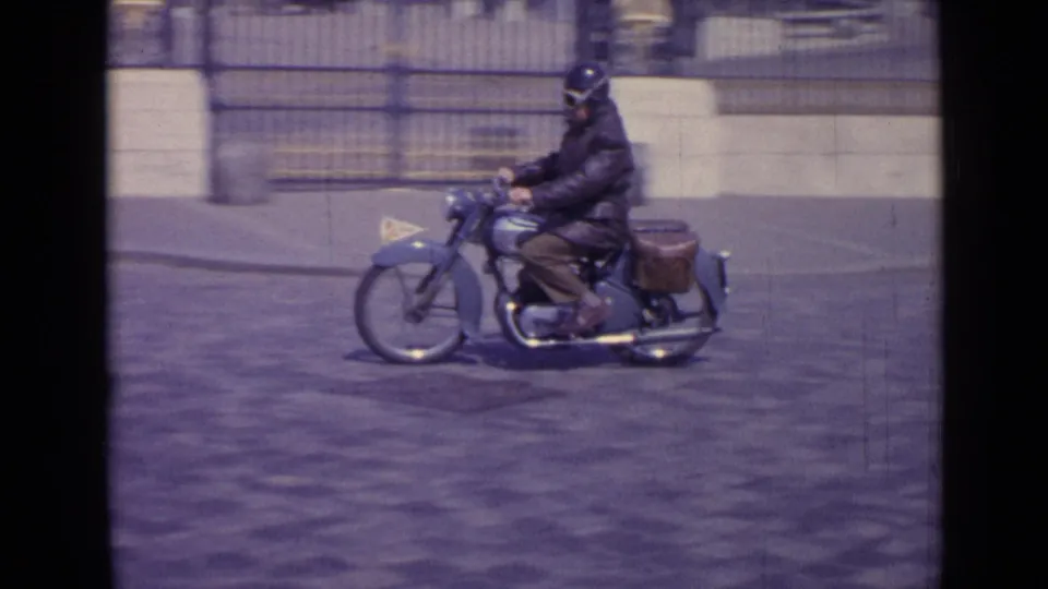 A helmeted motorcyclist with saddlebags rides past a stone building and iron fence in 1962 Versailles footage.