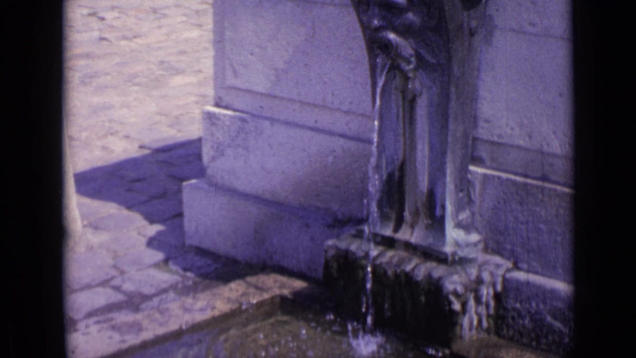 Stone fountain base with water falling into a shallow basin beside sunlit paving.