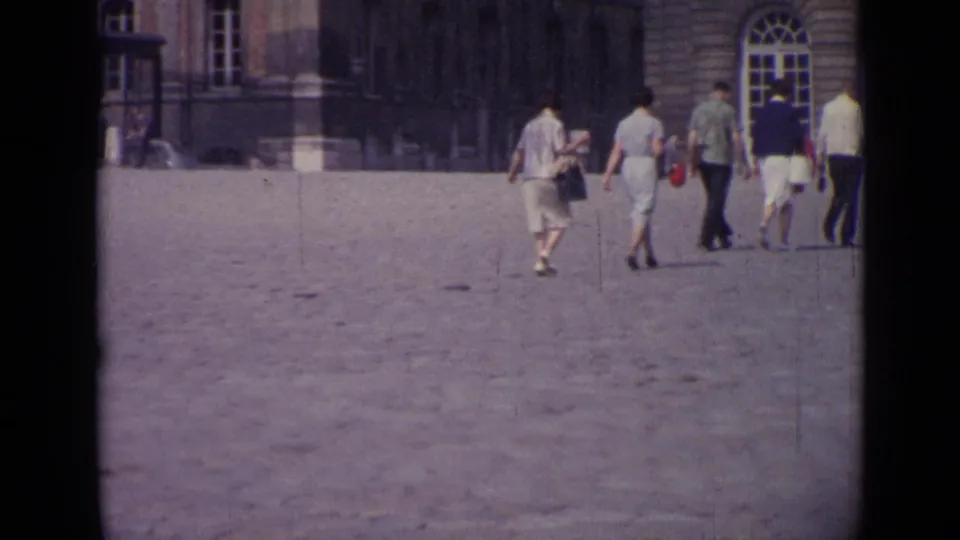 1962 Versailles frame showing tourists in mid-century clothing walking across the courtyard toward the palace facade.