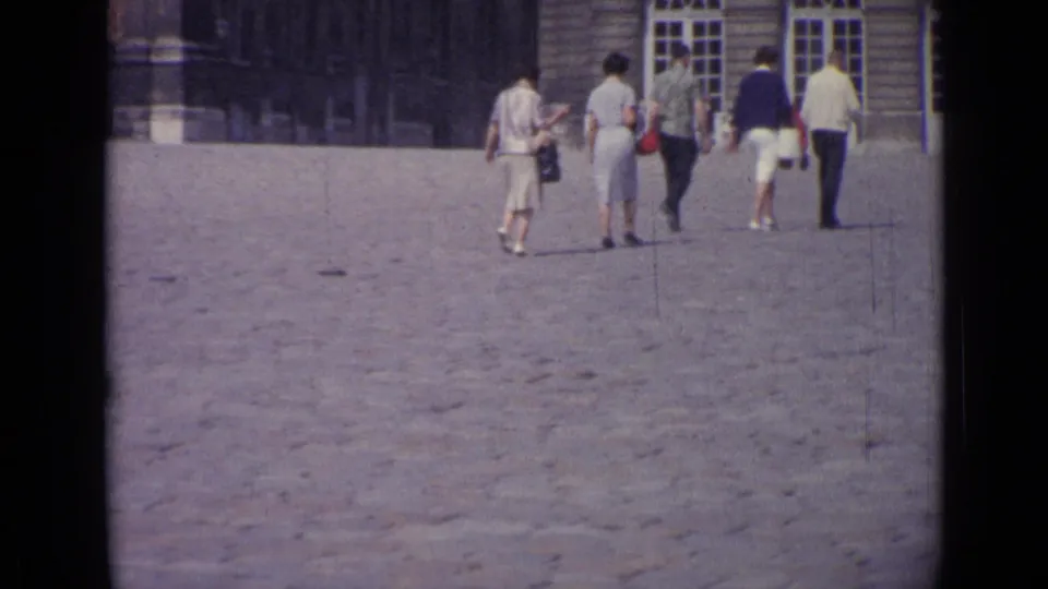 1962 Versailles home-movie frame with several tourists walking across the sunlit courtyard near palace windows.