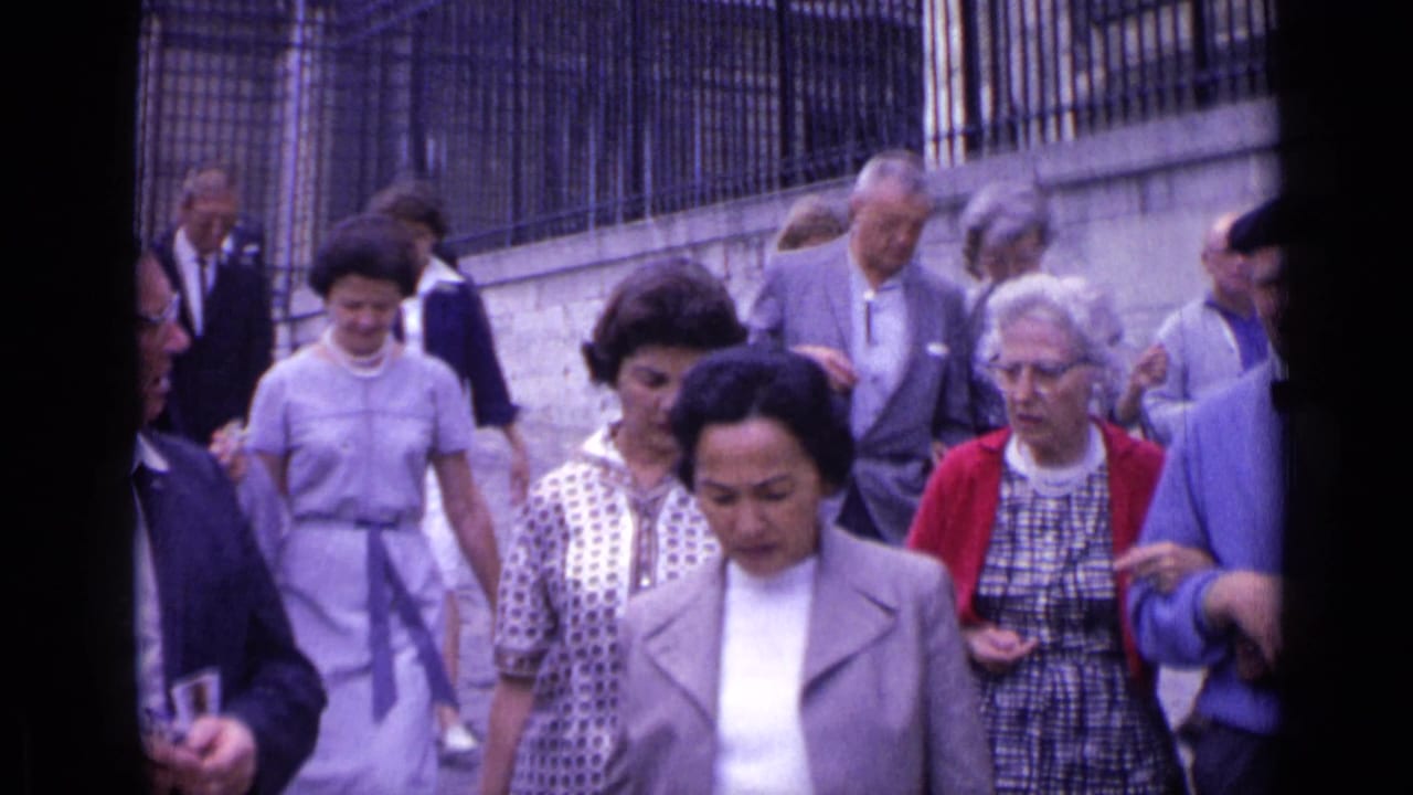 Tourists walk along a cobblestone street beside a stone wall and iron fence.