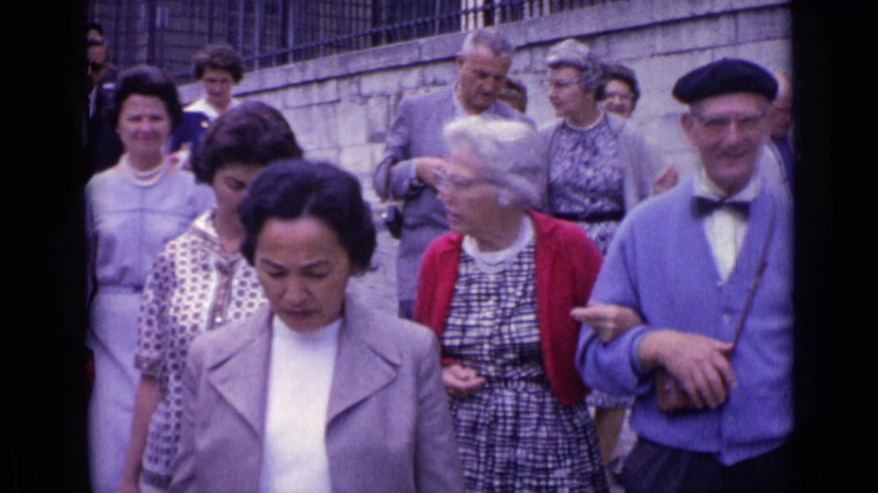 Adults walk downhill on cobblestones, including a woman in a light coat and a man in a beret.