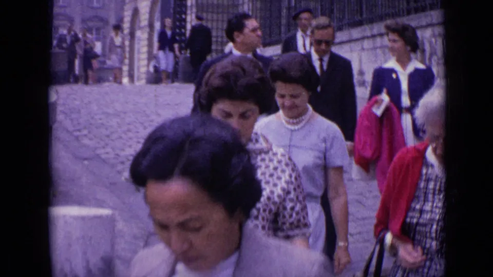 1962 Versailles frame showing adult tourists descending a cobblestone street, with background pedestrians, stonework, and formal clothing.