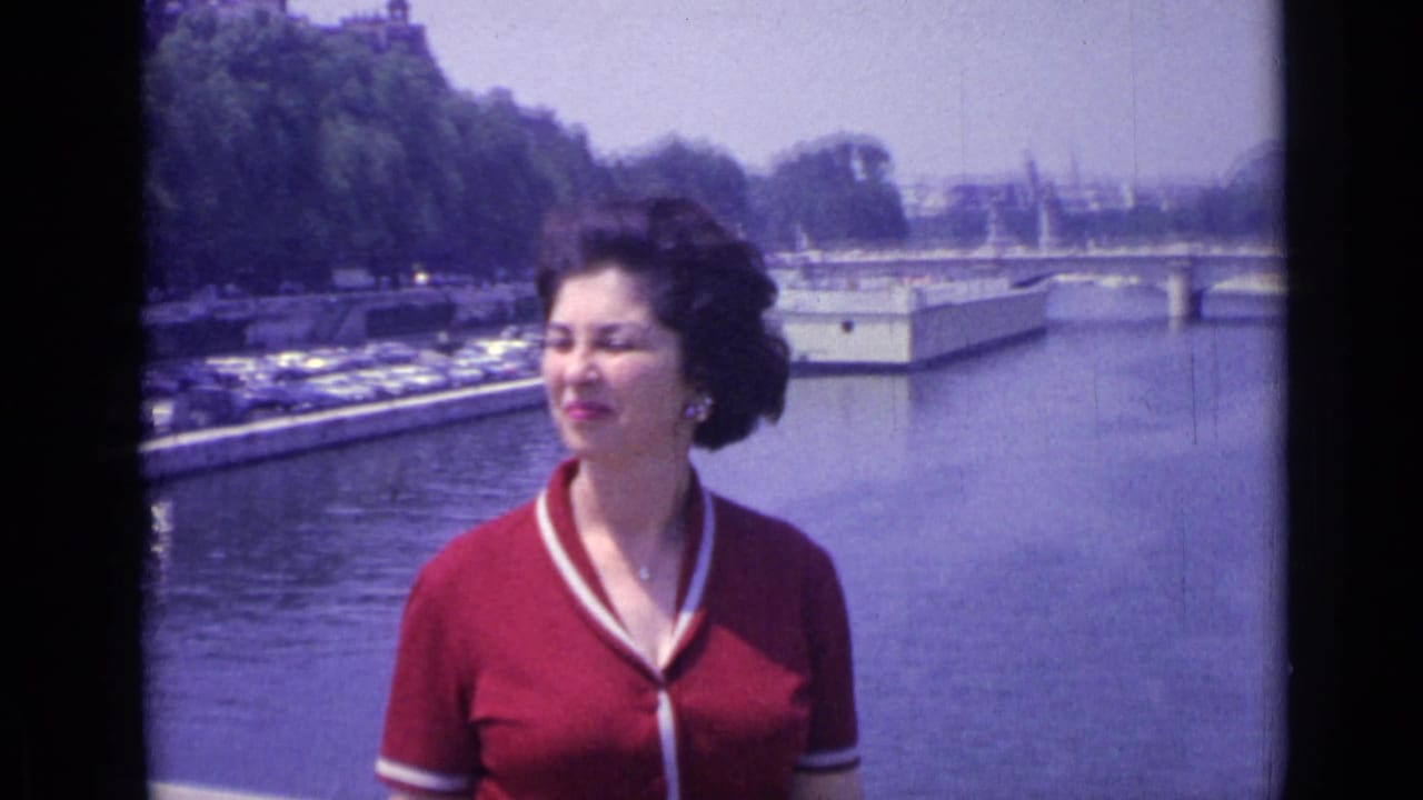 Woman in a red cardigan stands on a bridge with a river and parked cars behind her.
