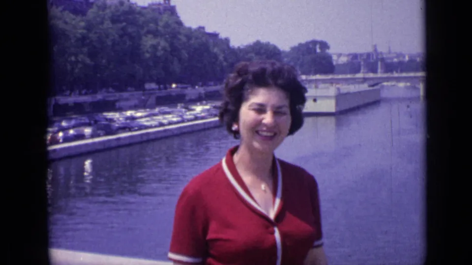 A smiling woman in 1962 Versailles stands on a bridge, framed by the river, trees, and parked cars behind her.