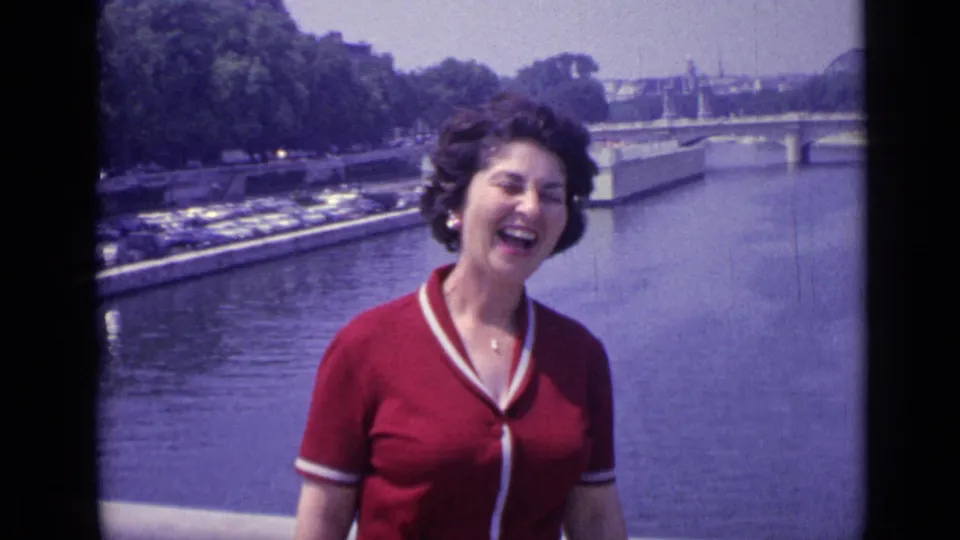 The woman laughs openly on a Versailles river bridge, with the water, parked cars, and trees visible behind her.