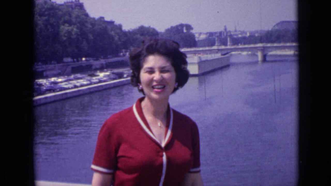 Woman in a red shirt faces the camera beside a river with a bridge behind her.