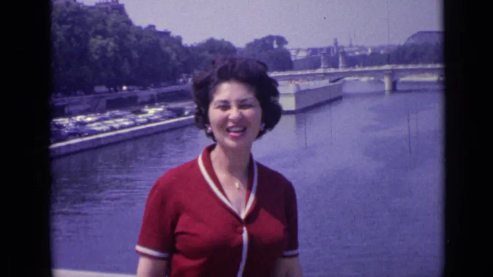 In Versailles, France in 1962, a woman in a red V-neck shirt smiles toward the camera beside a broad river and bridge.
