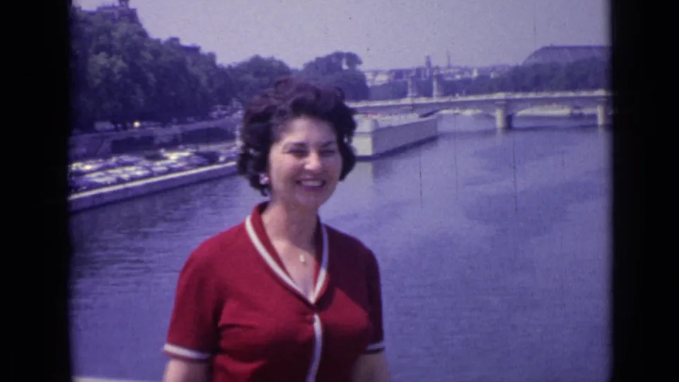 The woman turns slightly while smiling near the river in Versailles, with bridge spans and a tree-lined bank behind her.