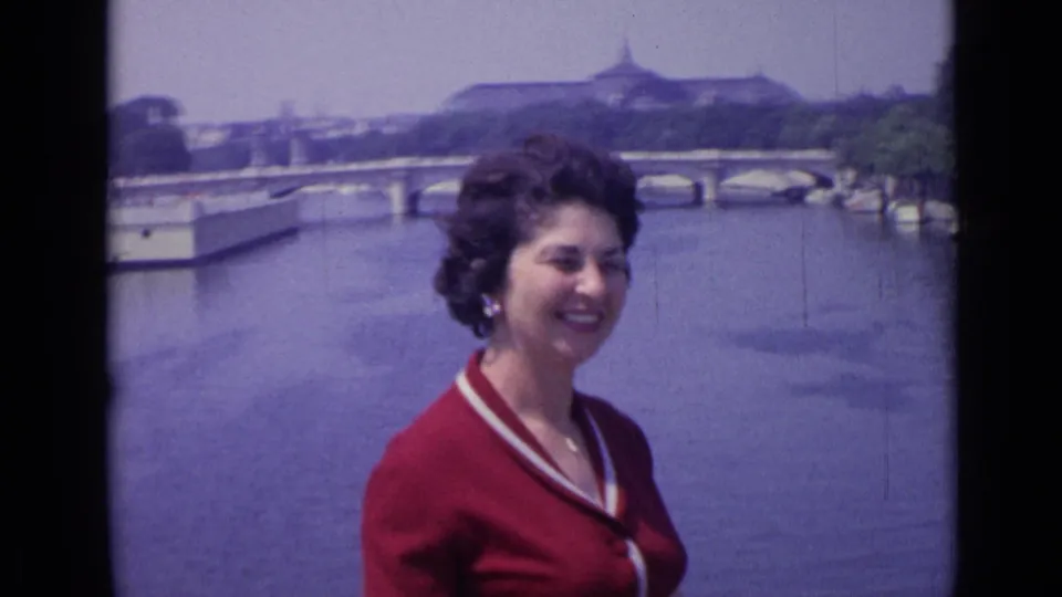 The woman smiles at the edge of the frame beside the Versailles river, with a bridge and distant large building visible across the water.