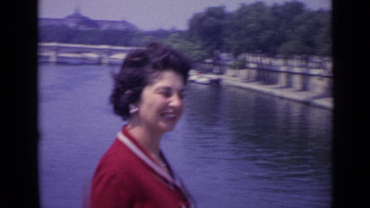 Woman in a red collared dress smiling beside a river with boats and a bridge behind her.