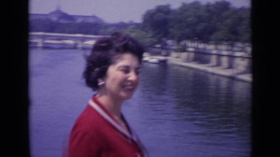 In 1962 France footage, a woman in a red dress smiles near a riverbank with small boats, trees, and a bridge in the background.