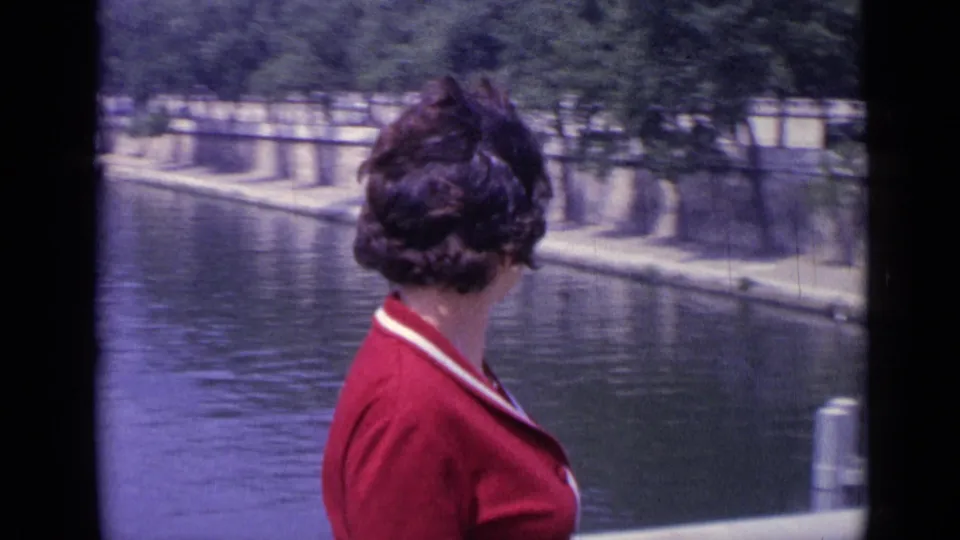 A woman in a red collared dress turns toward the water along a French riverbank, with trees and a paved embankment across the river.