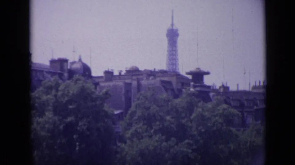 The Eiffel Tower appears in the distance above rooftops and leafy trees, filmed in soft 8mm color from a Paris-region vantage point.