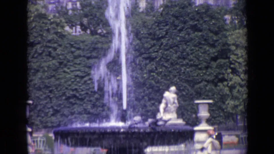 1962 Versailles garden frame showing a fountain spraying before dense greenery, with stone figures and a seated park visitor nearby.