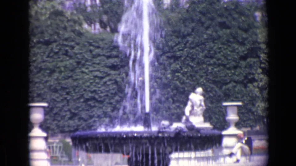 1962 Versailles home-movie frame with the fountain centered, water falling into the basin, stone garden ornaments, benches, and a seated visitor.