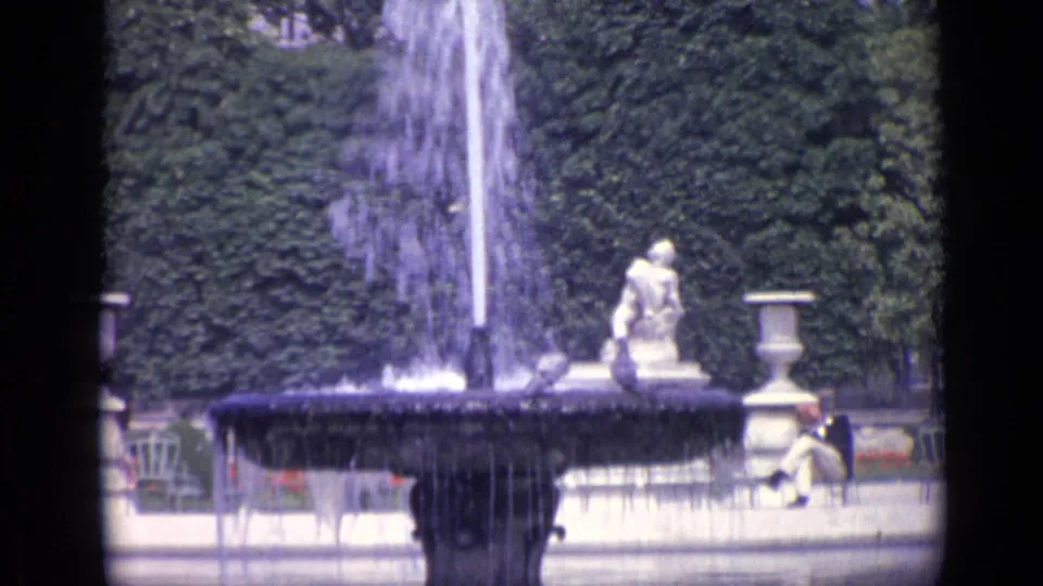 1962 Versailles garden frame showing a fountain and stone sculpture against dense hedges, with a seated person visible near the right edge.