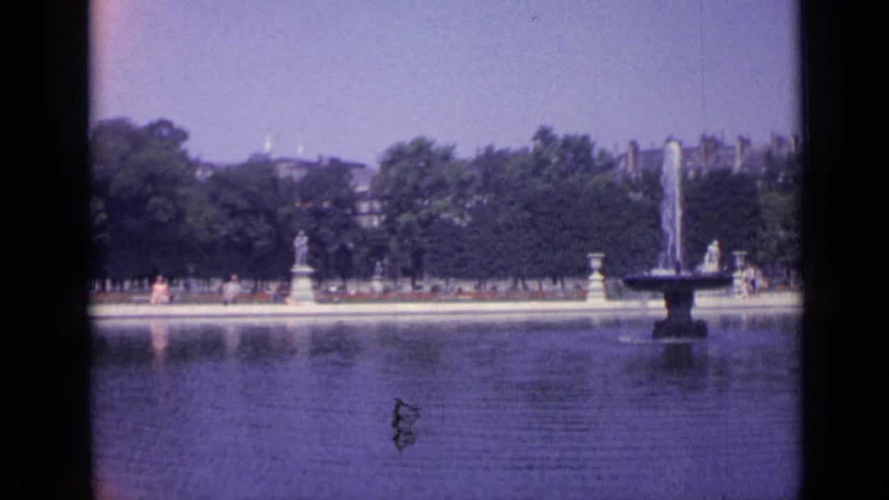 Wide 1962 Versailles view of the Grand Canal, fountain spray, formal garden edge, trees, statues, and distant visitors.