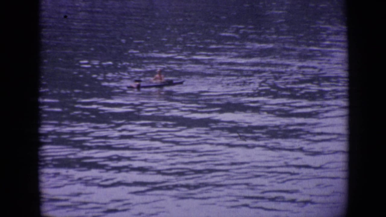 In Versailles, France in 1962, a distant person paddles a small canoe or kayak across calm rippled water in soft 8mm color.