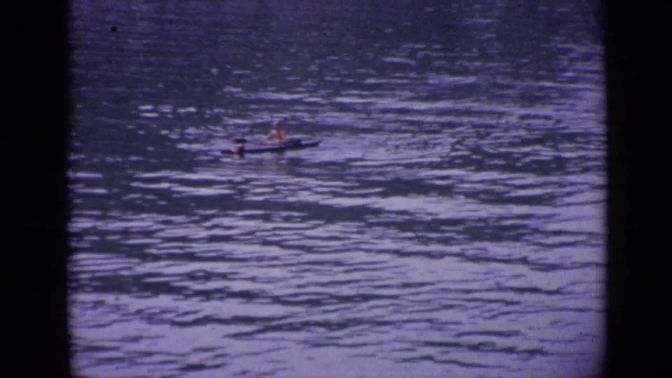 In Versailles, France in 1962, a distant person paddles a small canoe or kayak across calm rippled water in soft 8mm color.