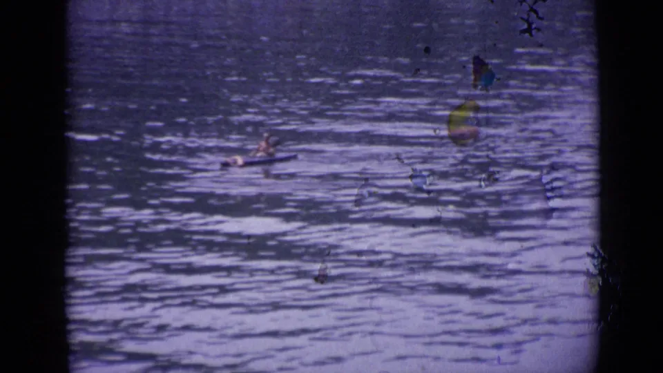 A distant paddler crosses the water in a small craft, with faint foliage at the right edge of the 1962 Versailles home-movie frame.