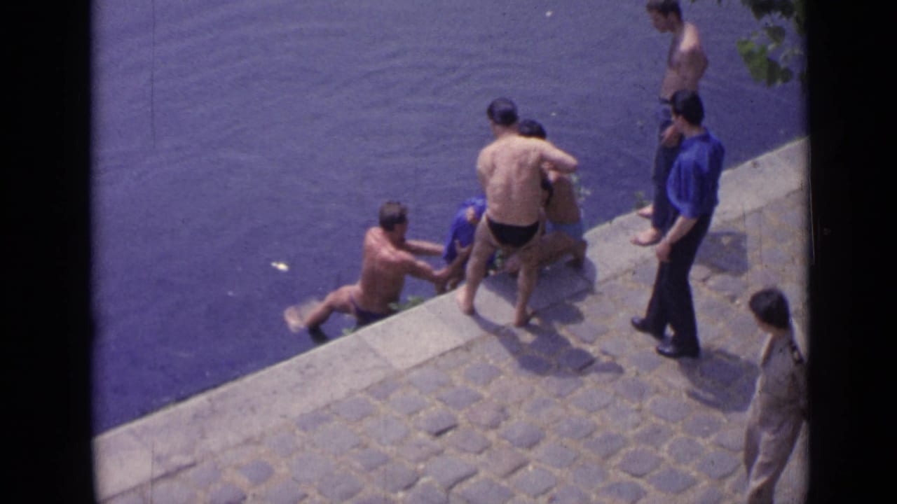 Several men reach toward a swimmer at the edge of a stone lakeside embankment.