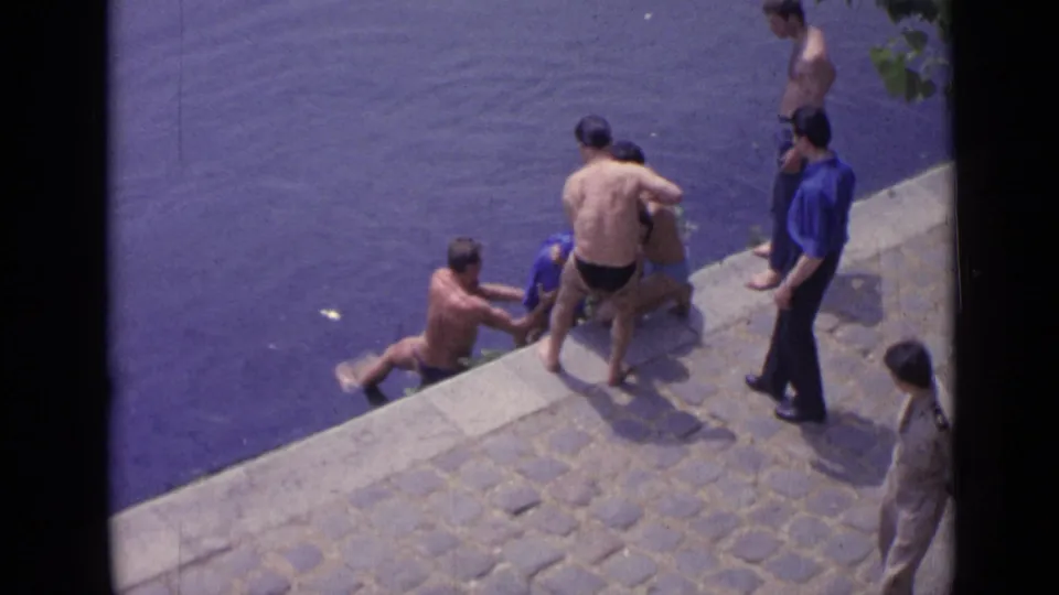 1962 Versailles footage frame with shirtless men and a man in a blue shirt helping a swimmer from the water.