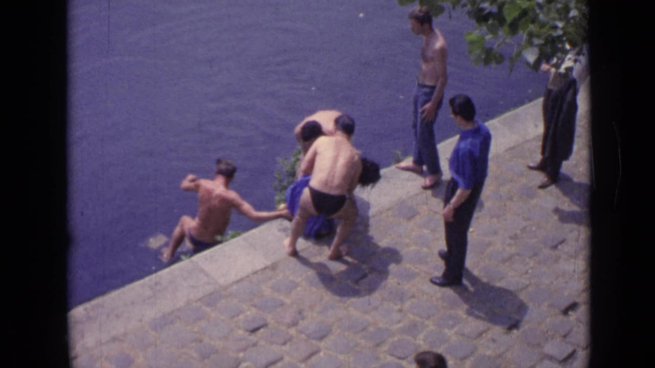 Men in swim trunks gather on a stone embankment beside the water.