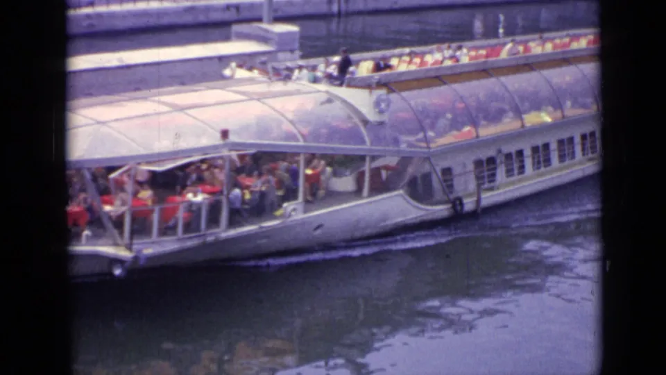 1962 Versailles home-movie frame showing a white canal tour boat with passengers under a curved glass roof and on an open upper deck.