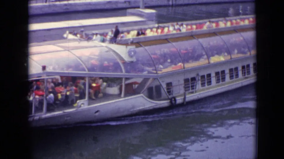 1962 Versailles frame of a white sightseeing boat passing by, with passengers visible through the transparent canopy and along the top deck.