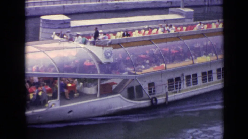 1962 Versailles home-movie frame with the tour boat closer in view, showing its curved roof, side windows, passengers, and canal setting.