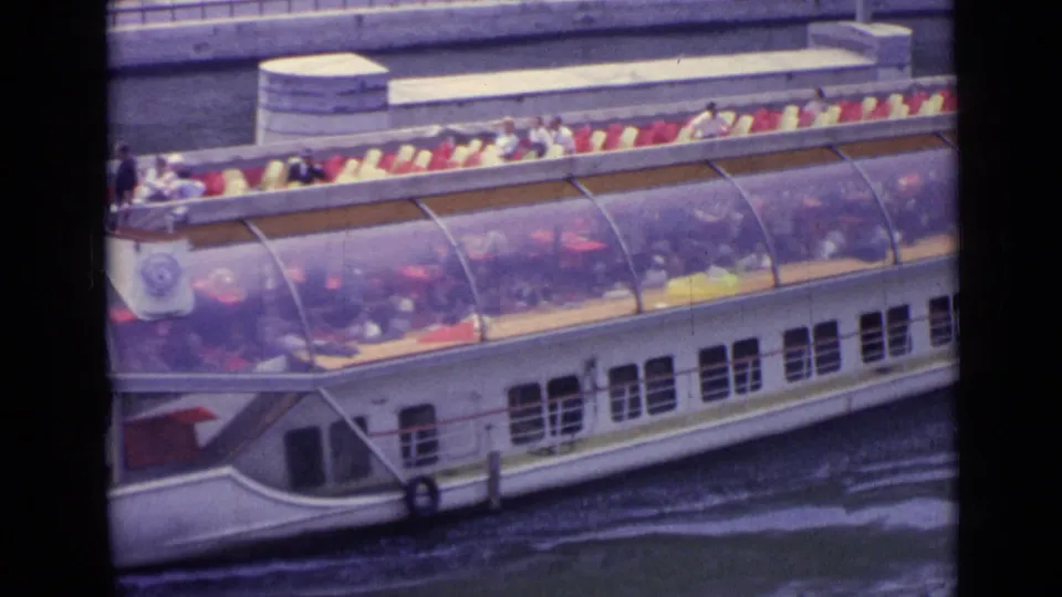 1962 Versailles frame showing the side of a white tour boat, its transparent roof, rows of passengers, and rippled canal water below.