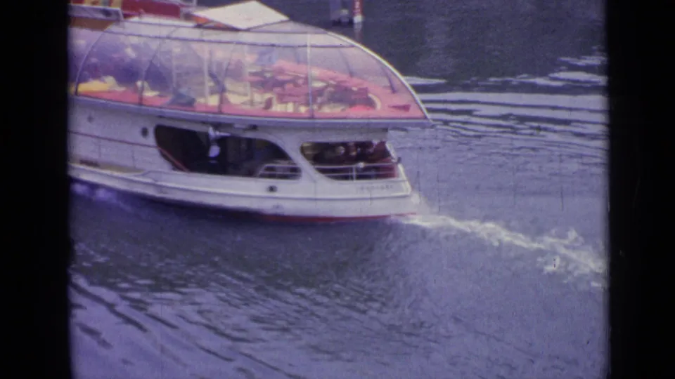 A closer side view of the glass-roofed river boat in Versailles, with its rear section and wake filling the frame.