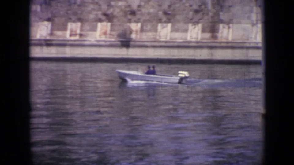 A small motorboat carrying two people moves along the river near a stone embankment in the 1962 Versailles footage.