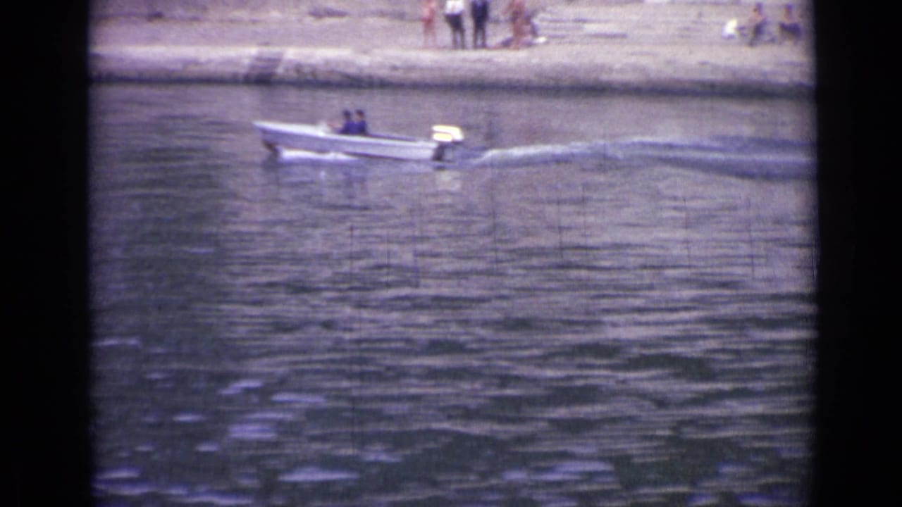 In 1962 France footage, a small white motorboat carries two people across a river near a sandy bank.
