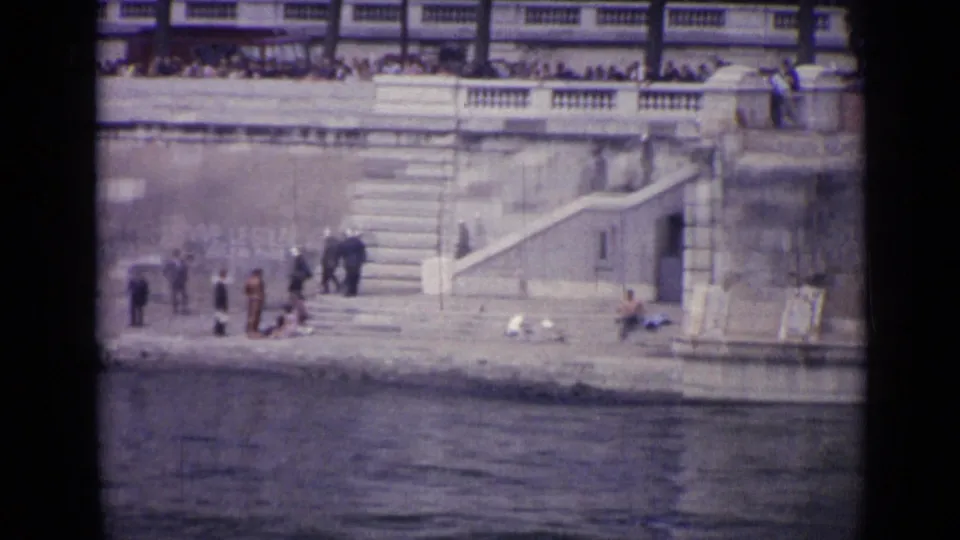 1962 Versailles home-movie frame showing people on a stone waterside embankment near stairs, with bystanders on the upper terrace.