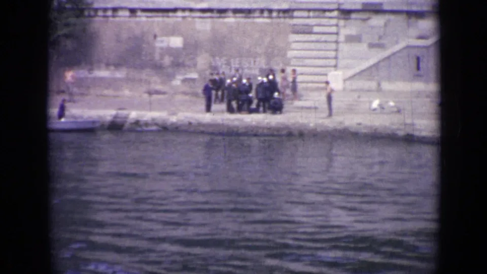 1962 Versailles frame showing a compact group of uniformed men near a stone staircase, with bystanders and water in the foreground.