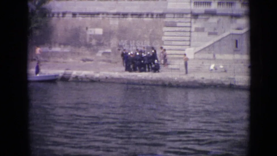 Distant 1962 Versailles frame of people standing along a concrete bank, with water, stone architecture, steps, and a small boat visible.