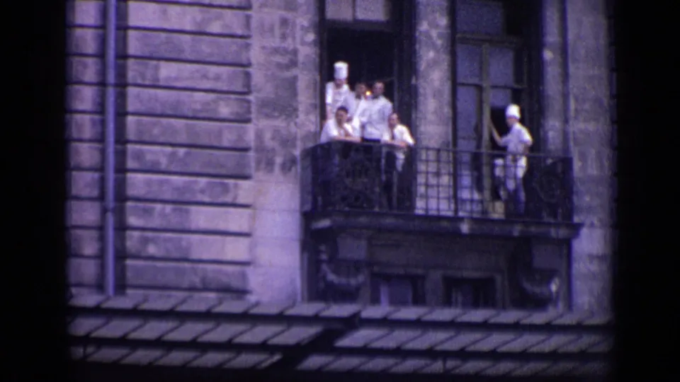 1962 Versailles frame showing chefs or kitchen staff in white uniforms and toques gathered on a wrought balcony of a stone building.