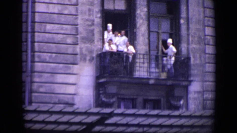 1962 Versailles home-movie frame of uniformed chefs on a balcony, framed by a stone wall, windows, and an open doorway.