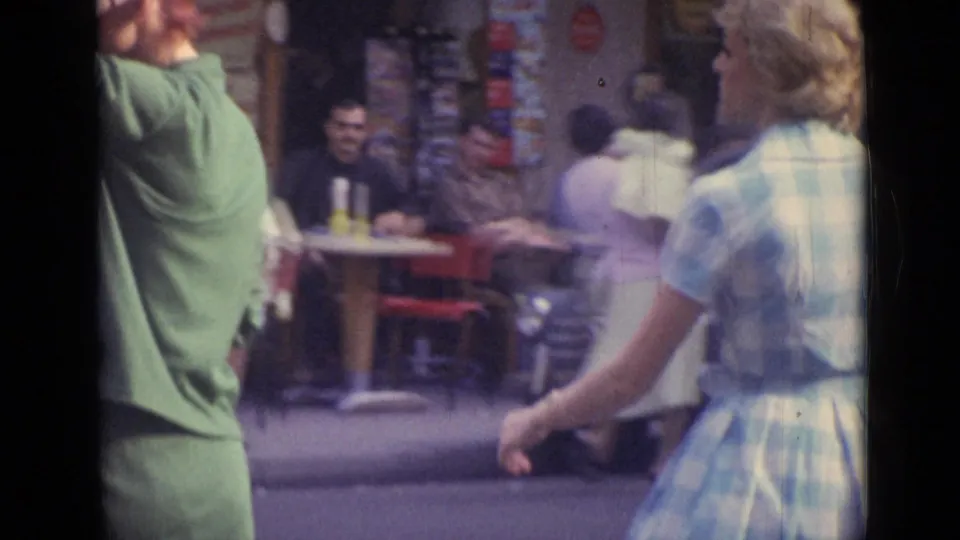 Women in 1960s dresses pass a sidewalk cafe area in Versailles, with seated patrons and shop displays behind them.