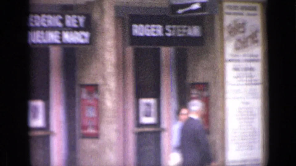 1962 Versailles theater frontage showing illuminated performer signs, poster cases, columns, and pedestrians in soft home-movie focus.