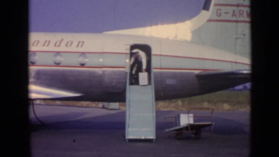 1962 airport frame showing a passenger descending mobile stairs from a Skyways of London aircraft, carrying or standing near a suitcase.