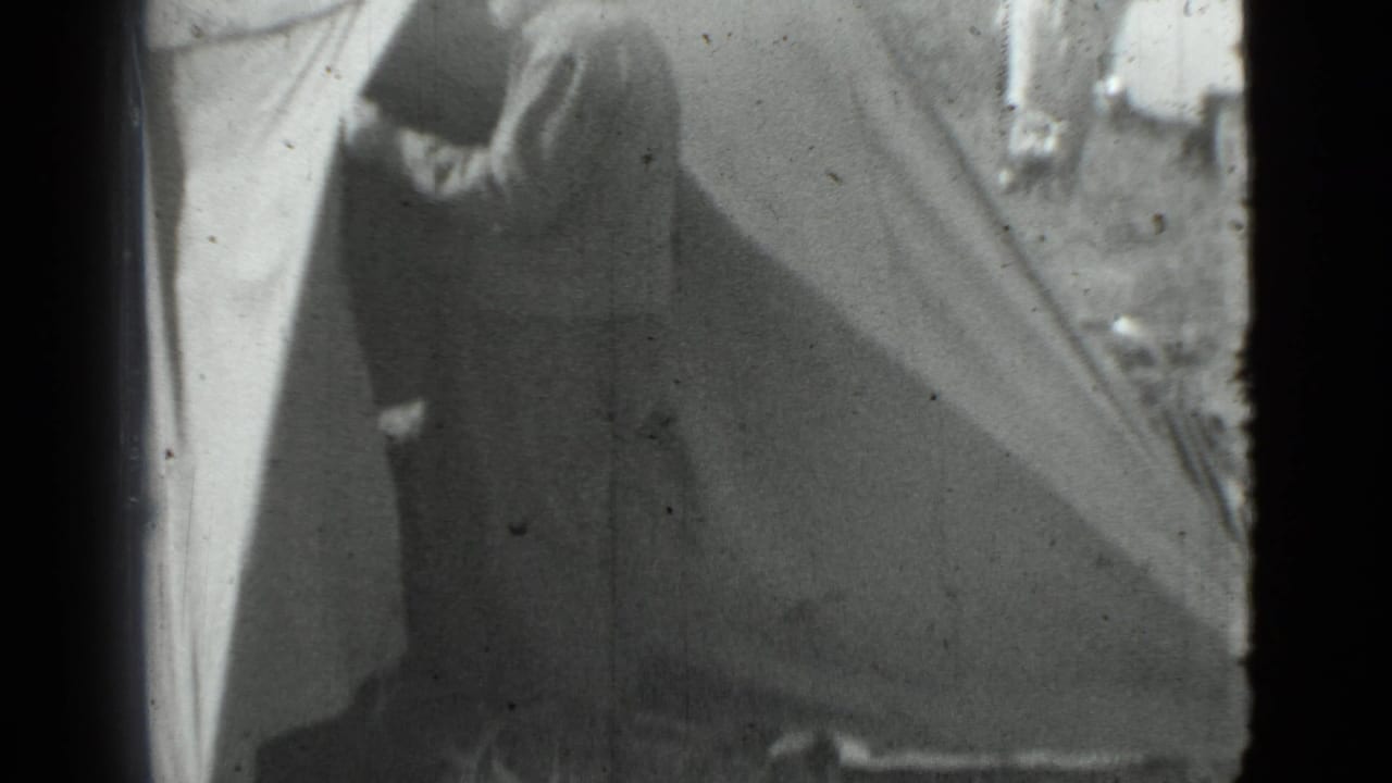 Man in a cap stands at the opening of a canvas tent, with rough ground and a car partly visible behind him.