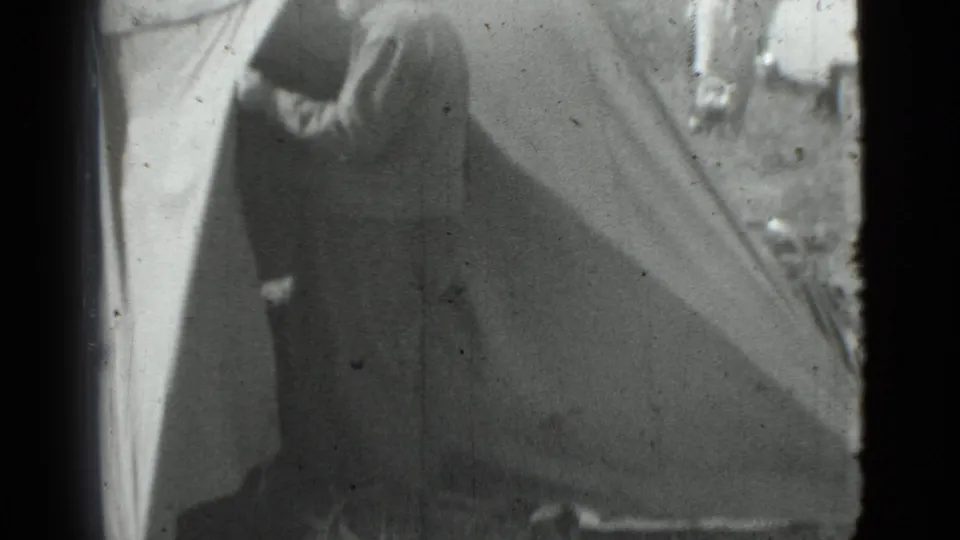 In supported 1967 Saint Petersburg home-movie footage, a man in a cap stands by a canvas tent entrance while background figures and a car appear nearby.