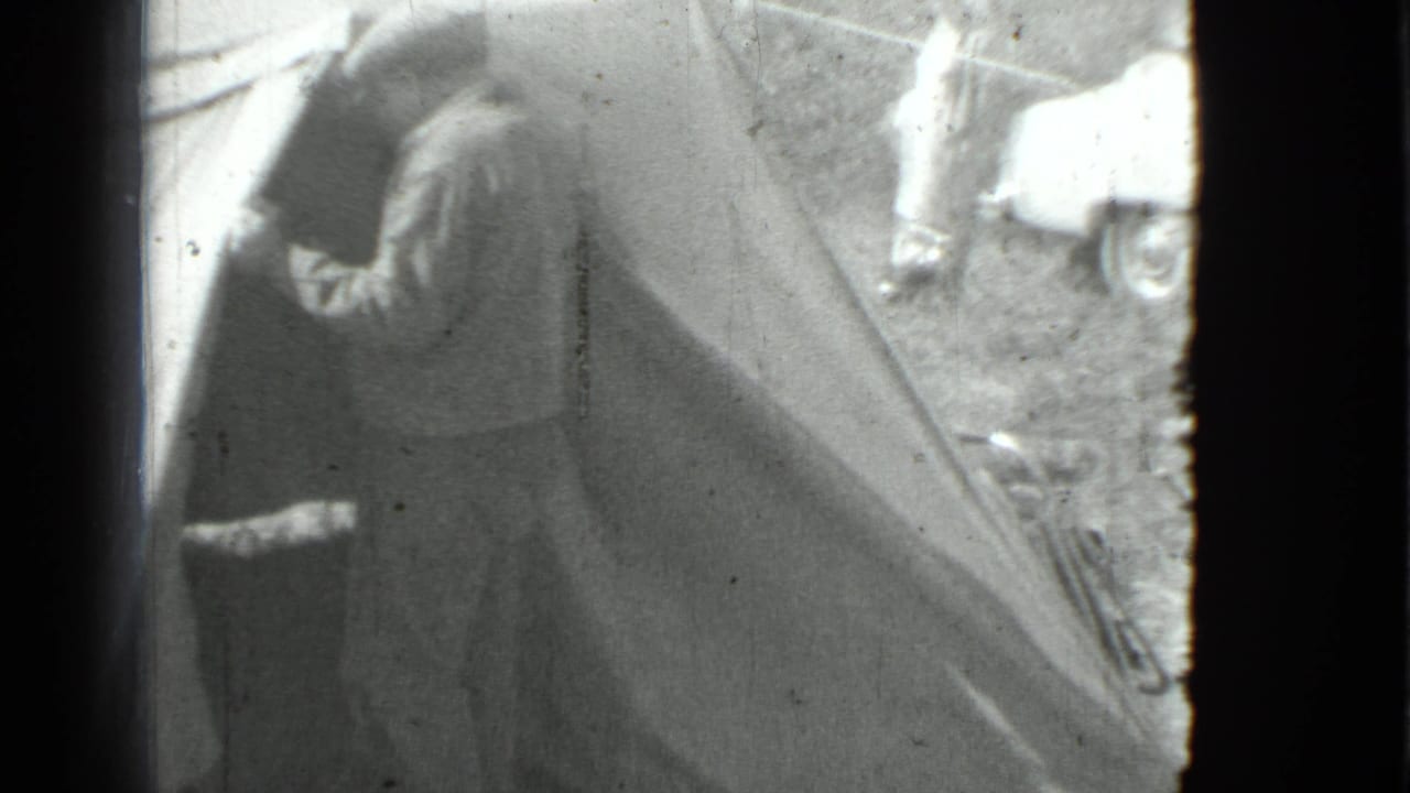 The man remains beside the tent entrance, appearing to look inside as a car and another person stand in the campsite background.