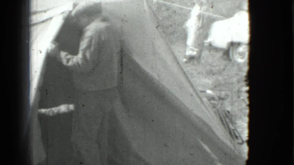 The man remains beside the tent entrance, appearing to look inside as a car and another person stand in the campsite background.